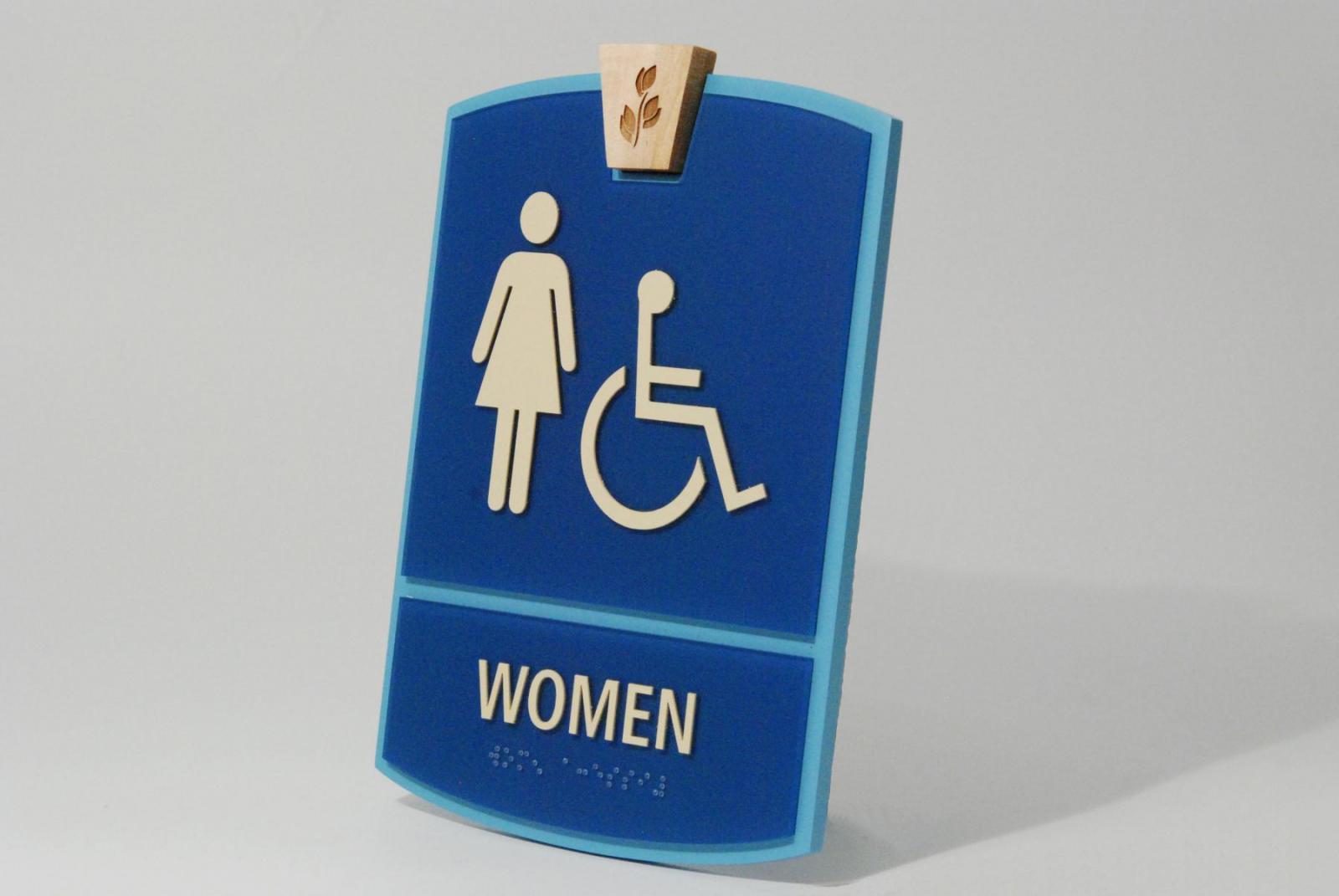 A blue restroom sign for women. It features white symbols of a standing woman and a person in a wheelchair. The word "WOMEN" is written in white letters at the bottom of the sign. A small wooden detail with leaves is above the symbols.