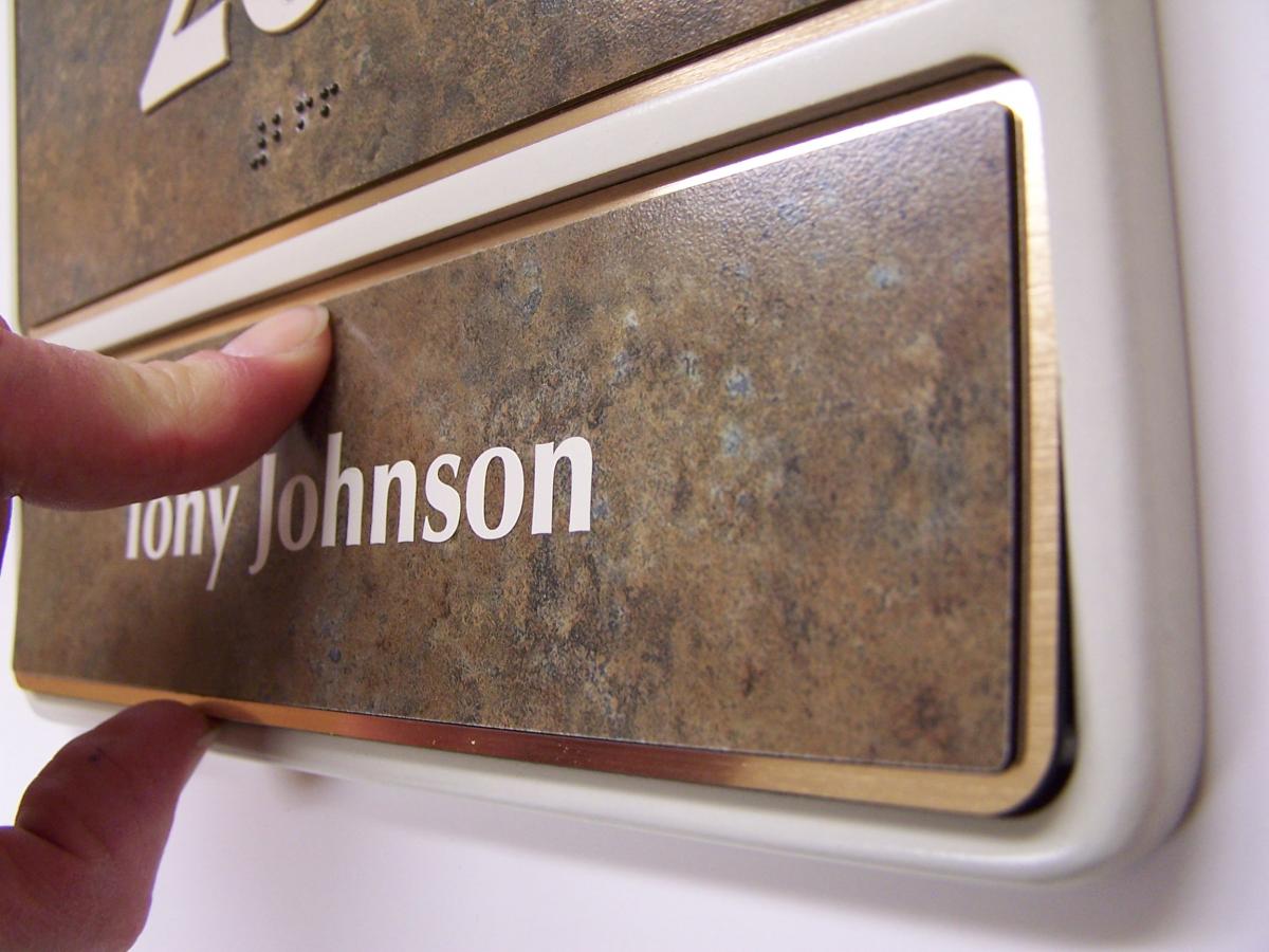 A close-up image of a nameplate with the text "Tony Johnson" in white letters on a textured brown background. A person's fingers are touching the bottom portion of the nameplate as if adjusting or removing it.