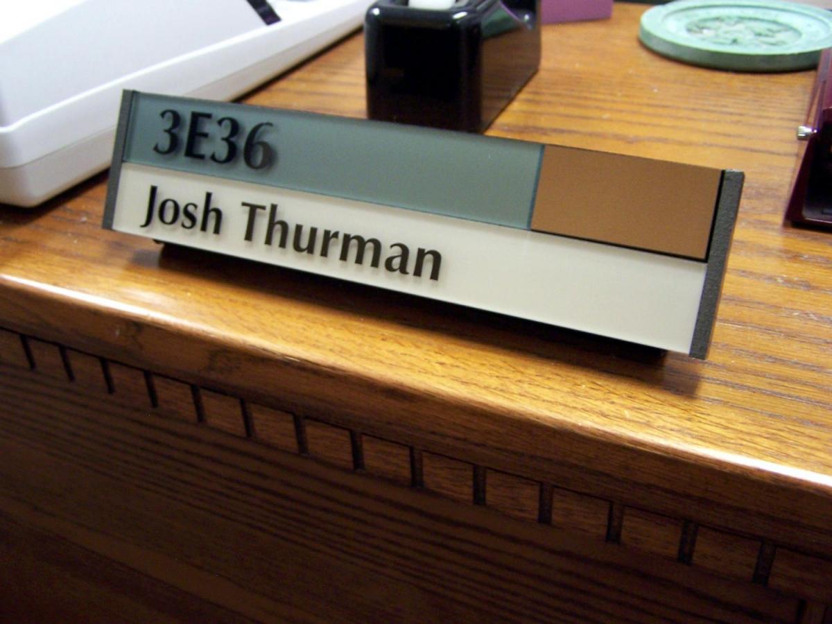 A desk nameplate displaying "3E36 Josh Thurman" in black text on a multi-colored background sits on a wooden desk. The nameplate has sections of blue, brown, and white. Various office items, including a computer keyboard, are partially visible in the background.