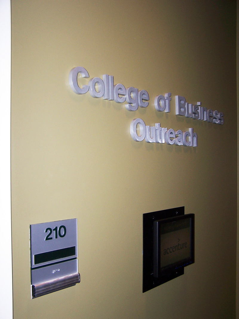 Signs on a beige wall indicate "College of Business Outreach." Additional sign shows a room labeled 210, featuring a display screen beneath the sign.