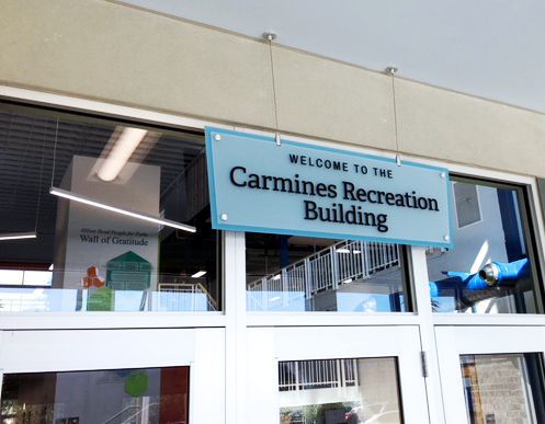 A light blue sign with white lettering reads "Welcome to the Carmines Recreation Building" and is mounted above the entrance doors of a building. Inside, there's a blurred wall with a sign reading "Wall of Gratitude." The interior appears well-lit and modern.