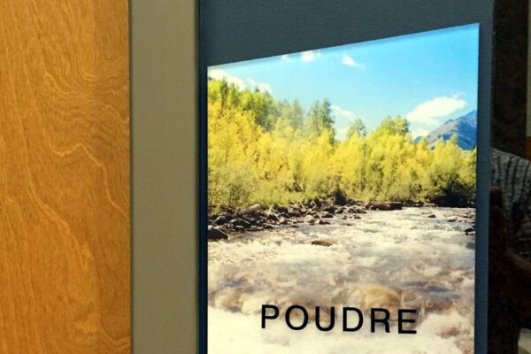 A sign with an image of a river and trees under a bright blue sky with some clouds. The word "POUDRE" is written over the image. The sign is mounted on a wall next to a wooden door.