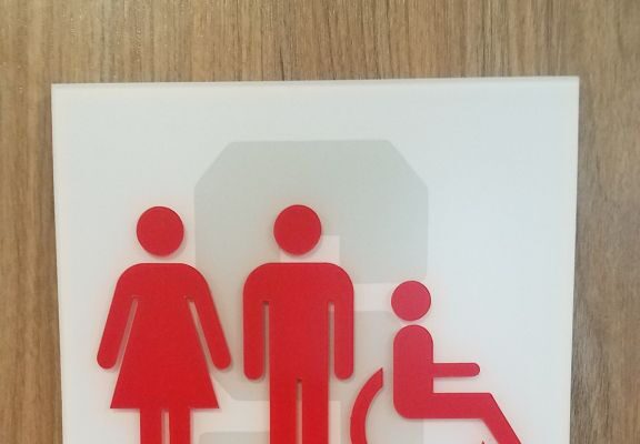 A family restroom sign on a wooden surface, featuring icons in red. From left to right, the icons depict a woman, a man, and a person using a wheelchair. The text "FAMILY RESTROOM" is written below the icons. Braille is present beneath the text.