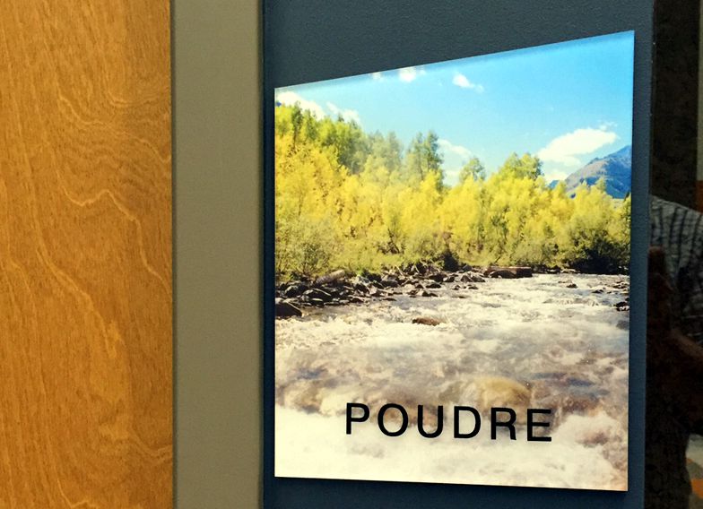 A sign with an image of a river and trees under a bright blue sky with some clouds. The word "POUDRE" is written over the image. The sign is mounted on a wall next to a wooden door.