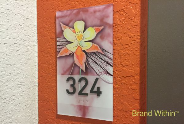 A decorative sign on an orange wall displays the number "324" below an illustration of a flower with white petals and orange accents. The textured wall beside it is white. In the lower right corner, the text "Brand Within®" is visible.