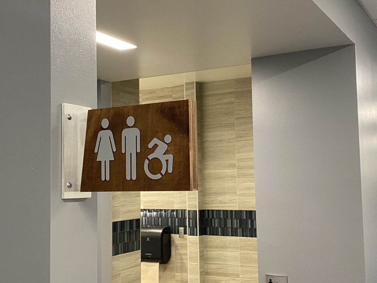 A restroom sign mounted on a light-colored tiled wall depicts icons for male and female restrooms and an additional icon indicating wheelchair accessibility.