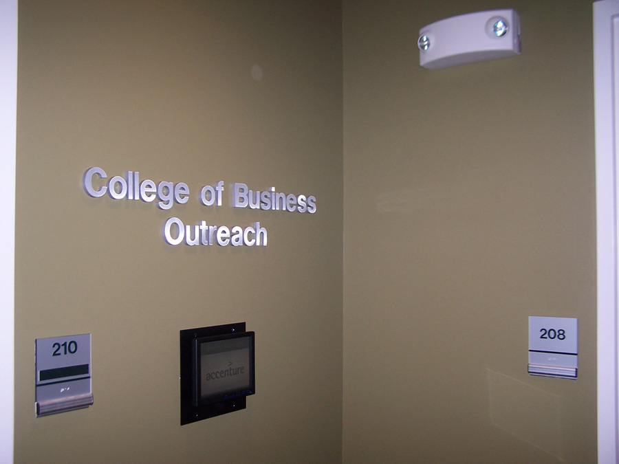 Signs on a beige wall indicate "College of Business Outreach." Additional signs show a room labeled 210, featuring a display screen beneath the sign, and another room labeled 208. A ceiling light fixture is visible above.