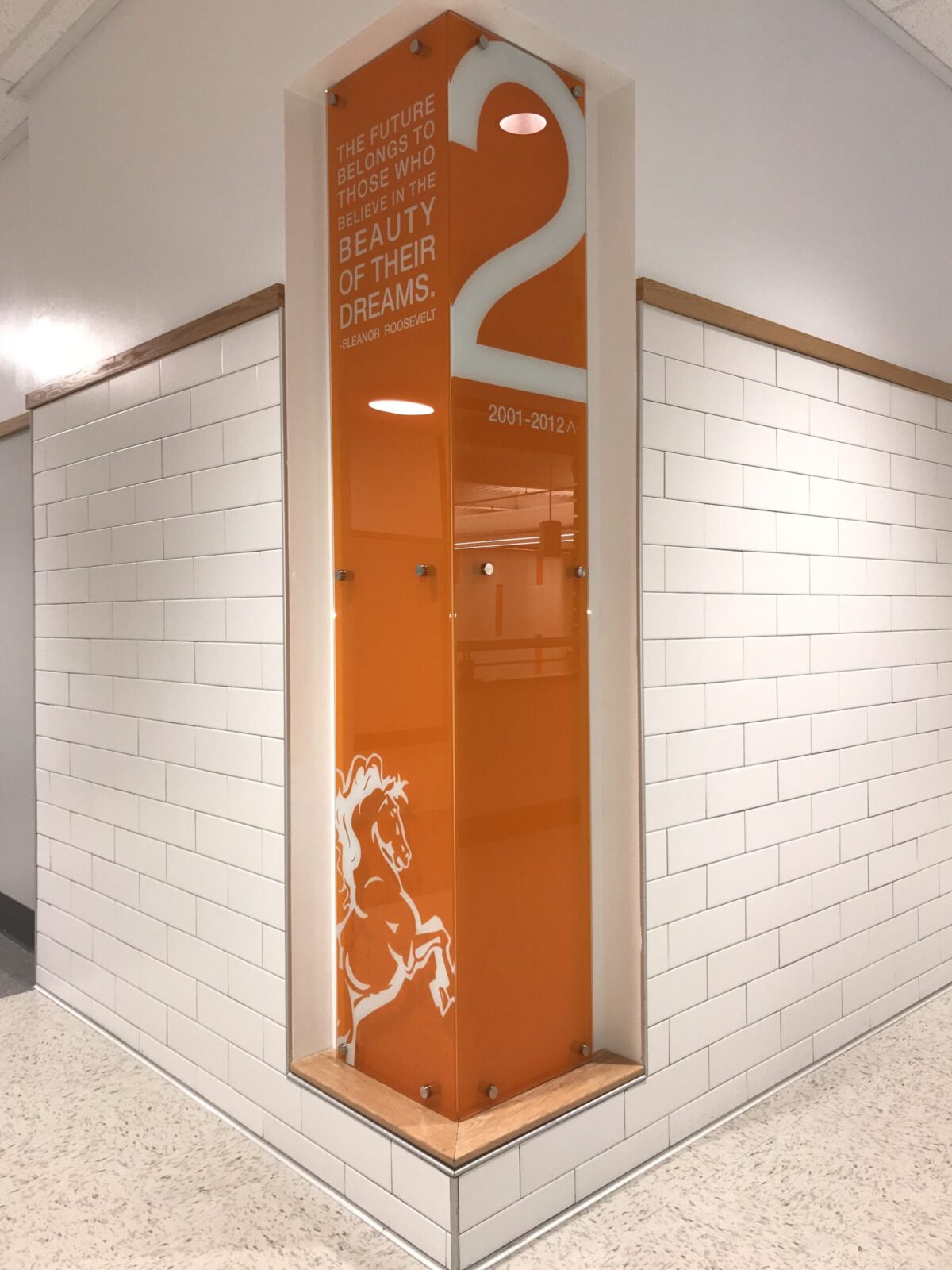 An interior hallway corner featuring a tall orange display with the number "2" and the name "John Bell." The display has a quote about dreams and an illustration of a horse in motion at the bottom. The wall is tiled in white bricks.