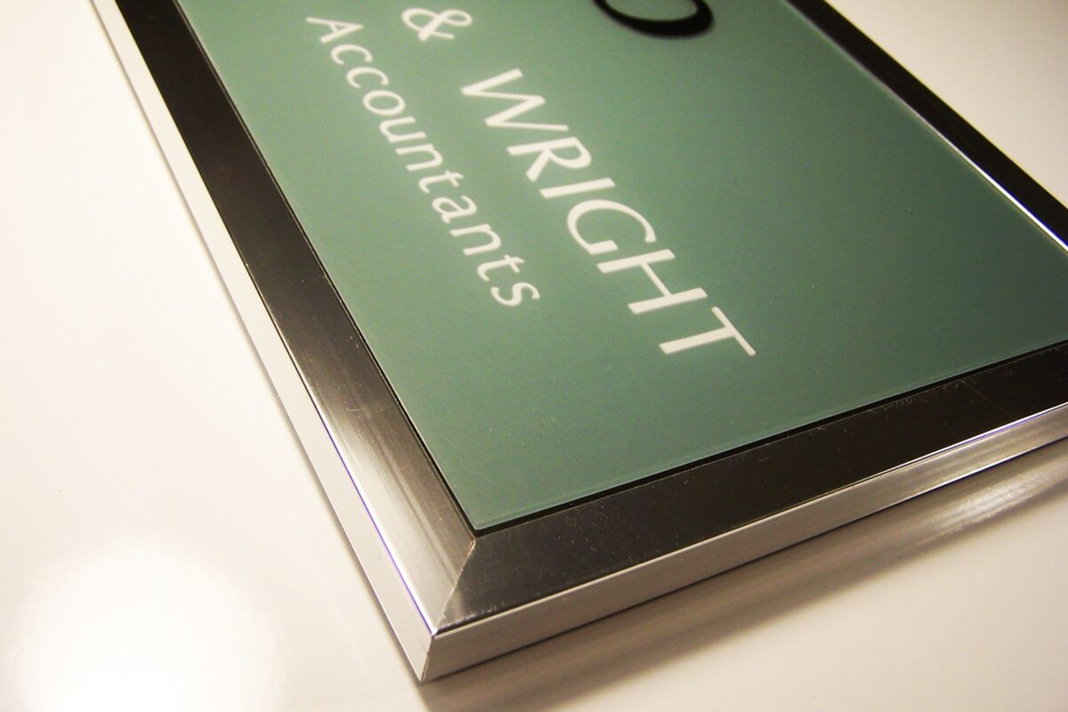 A close-up image of a rectangular, metallic-framed sign that reads "RIGHT & WRIGHT Accountants" against a green background. The sign has a sleek and polished appearance, with the text concentrated more towards the right side of the frame.