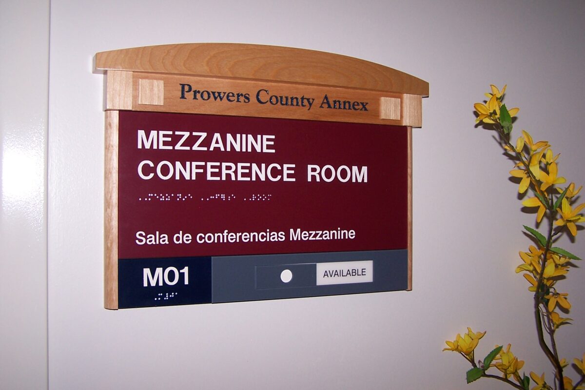 A sign on a wall reads "Prowers County Annex" at the top. Below it, the sign indicates "Mezzanine Conference Room" and "Sala de conferencias Mezzanine," with Braille text as well. The sign is marked with room number "M01" and a label showing availability status. Yellow flowers are visible to the right.