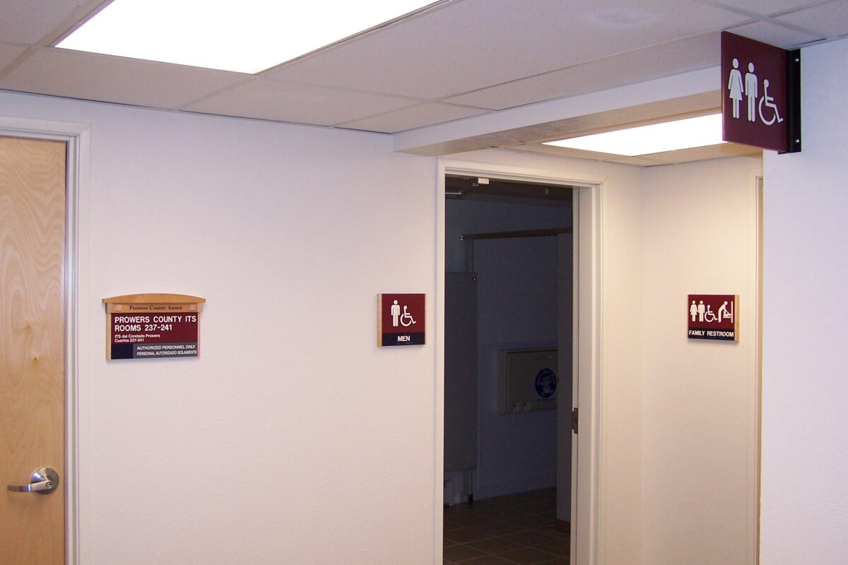 A hallway features a partially open door to a restroom with a red sign displaying male and female figures and a wheelchair symbol. Adjacent to the door, another sign reads "MEN." On the wall beside the door, a plaque is visible.