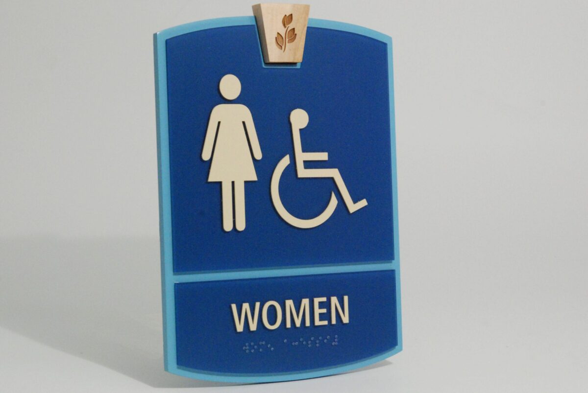 A blue restroom sign for women. It features white symbols of a standing woman and a person in a wheelchair. The word "WOMEN" is written in white letters at the bottom of the sign. A small wooden detail with leaves is above the symbols.