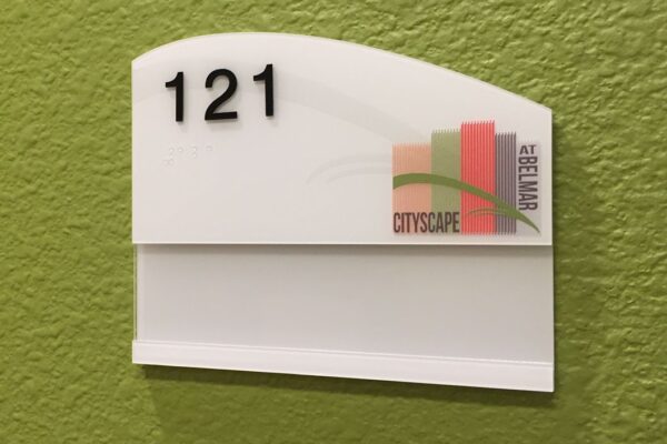 A green wall with a white, rectangular room number sign showing "121" in black text at the top. Below the number, there's a multicolored logo with abstract building shapes and the words “CITYSCAPE” on the right side.