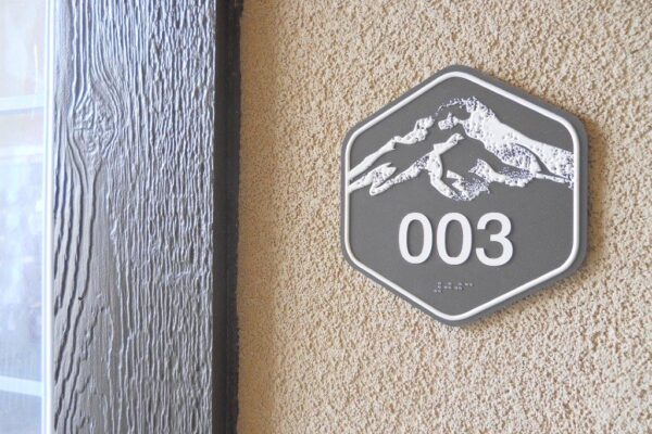 A hexagonal sign on a textured beige wall shows the number "003" with a white mountain graphic above it. To the left, a doorframe with a wood grain pattern is partially visible.