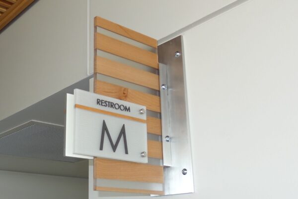 A restroom sign mounted on a wall indicates the men's restroom with a letter "M". The sign is attached to a wooden panel with horizontal slats. The word "RESTROOM" appears above the "M" on the sign.