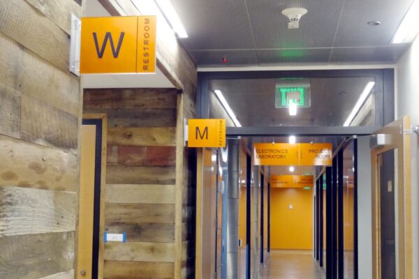 A hallway with wood-paneled walls and orange signs indicating restrooms. One sign with a 'W' for women's restroom is near a door on the left, and another sign with an 'M' for men's restroom is further down the hall. The ceiling has fluorescent lights and an exit sign.