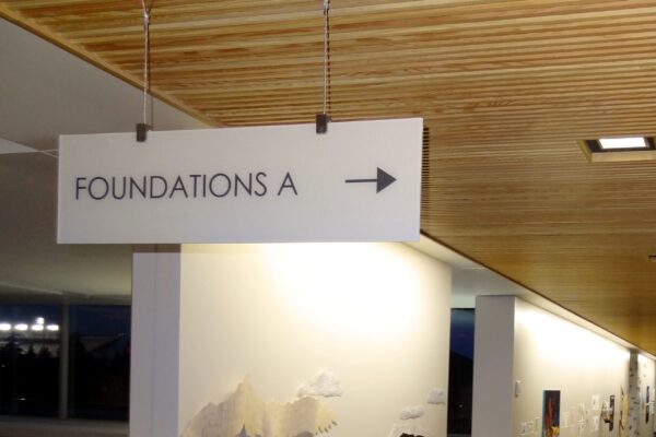 A ceiling-mounted sign in a hallway with a wooden slat ceiling points to the right. The sign reads "FOUNDATIONS A" in capital letters. The hallway has textured wall artworks and soft lighting, creating a modern and inviting atmosphere.