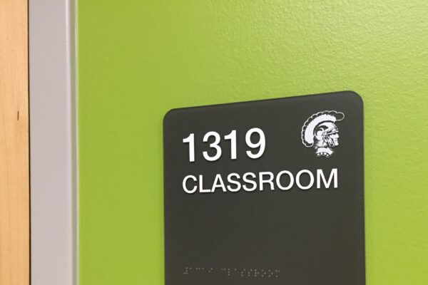 A green wall with a black sign displaying the room number "1319" in white text, above the word "CLASSROOM." The sign also includes a small image of a gladiator helmet and has Braille text at the bottom.