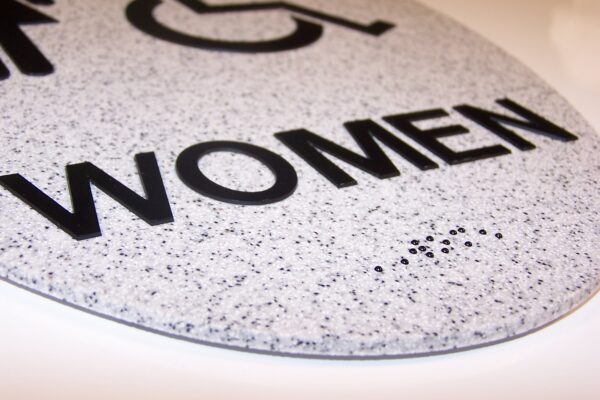 A close-up view of a gray restroom sign with black, embossed letters that spell out "WOMEN" and a wheelchair accessibility symbol above it. The surface appears textured with small, darker specks, providing a tactile quality.